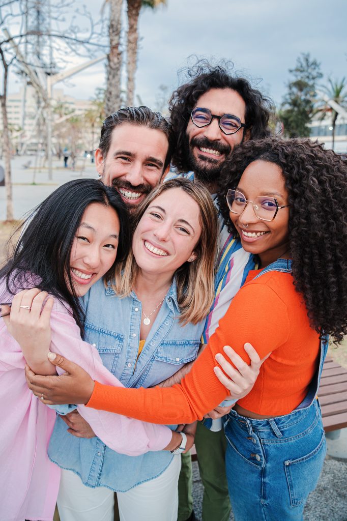 Vertical portrait of group of joyful young adult friends hugging each others. Happy smiling multiracial people embracing and laughing together on a social gathering. Community, union and friendship. High quality photo